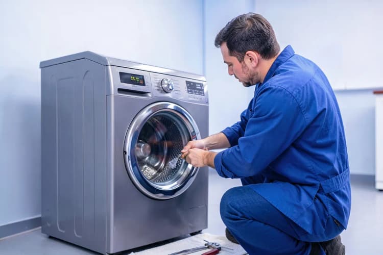 Um técnico de uniforme azul consertando uma máquina de lavar e secar roupa LG cinza, com ferramentas sobre um pano no chão.