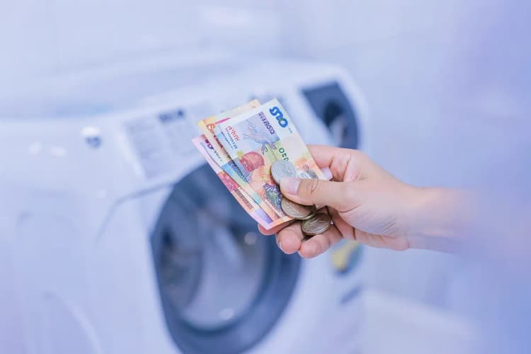 Uma mão segurando notas de dinheiro e moedas ao lado de uma máquina de lavar e secar roupa.