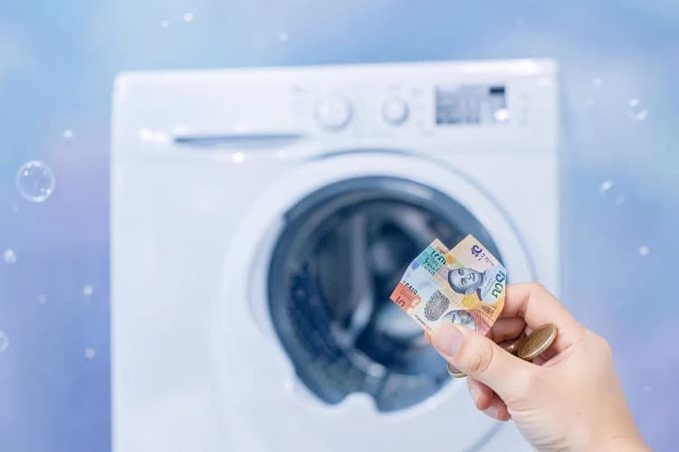 Mão segurando notas de dinheiro e moedas ao lado de uma máquina de lavar roupa, plano detalhe, luz de teto, dia