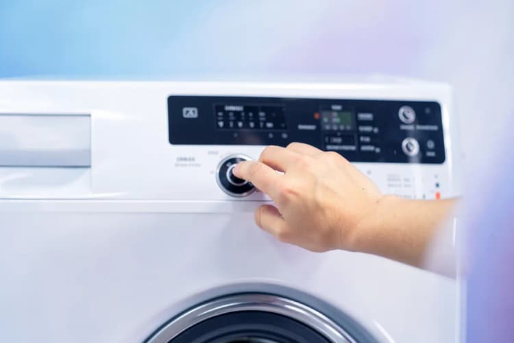Mão humana pressionando o botão de ligar de uma máquina lava e seca Samsung com o painel apagado.
