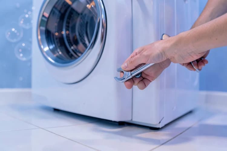 Pés niveladores de uma máquina de lavar e secar sendo ajustados com uma chave inglesa.