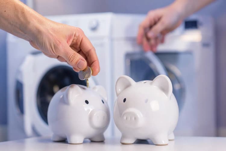 Mão colocando moedas em um cofrinho ao lado de uma máquina de lavar e secar.