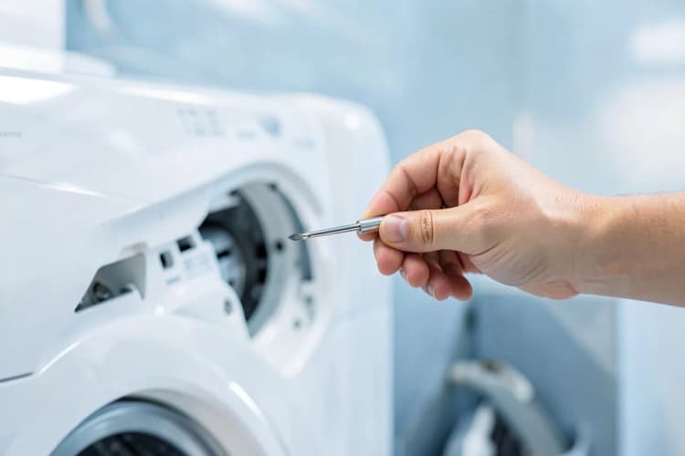 Uma mão humana segurando uma pequena chave de fenda, perto de um painel interno de máquina de lavar.